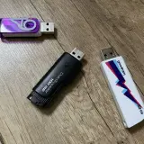 Trzy pendrive'y na drewnianym tle. Jeden fioletowy z logo, czarny DataTraveler 100 GB i biały Goodram 32 GB. Szybki pendrive 64 GB to marzenie!