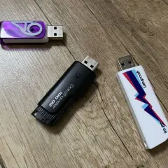 Trzy pendrive'y na drewnianym tle. Jeden fioletowy z logo, czarny DataTraveler 100 GB i biały Goodram 32 GB. Szybki pendrive 64 GB to marzenie!