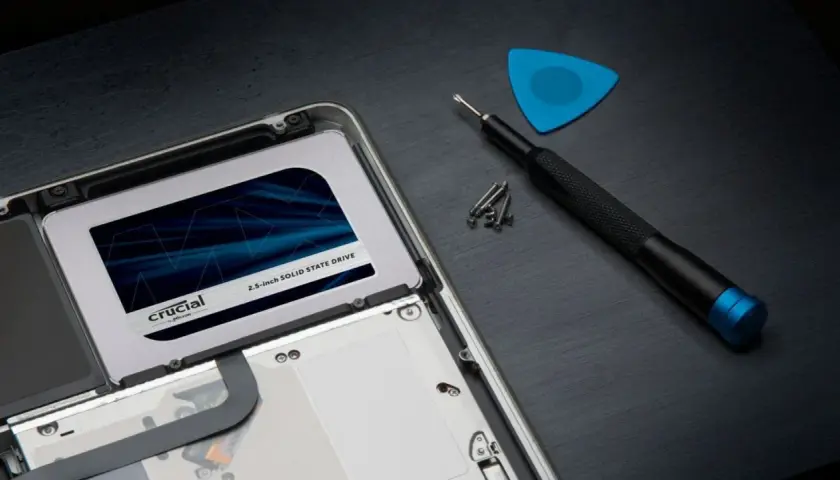 Wymiana dysku na **Crucial MX500 4TB** w laptopie. Obok śrubokręt, śrubki i niebieski pick.