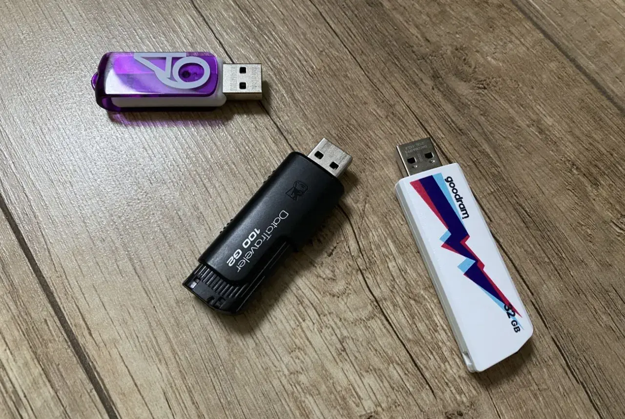 Trzy pendrive'y na drewnianym tle. Jeden fioletowy z logo, czarny DataTraveler 100 GB i biały Goodram 32 GB. Szybki pendrive 64 GB to marzenie!