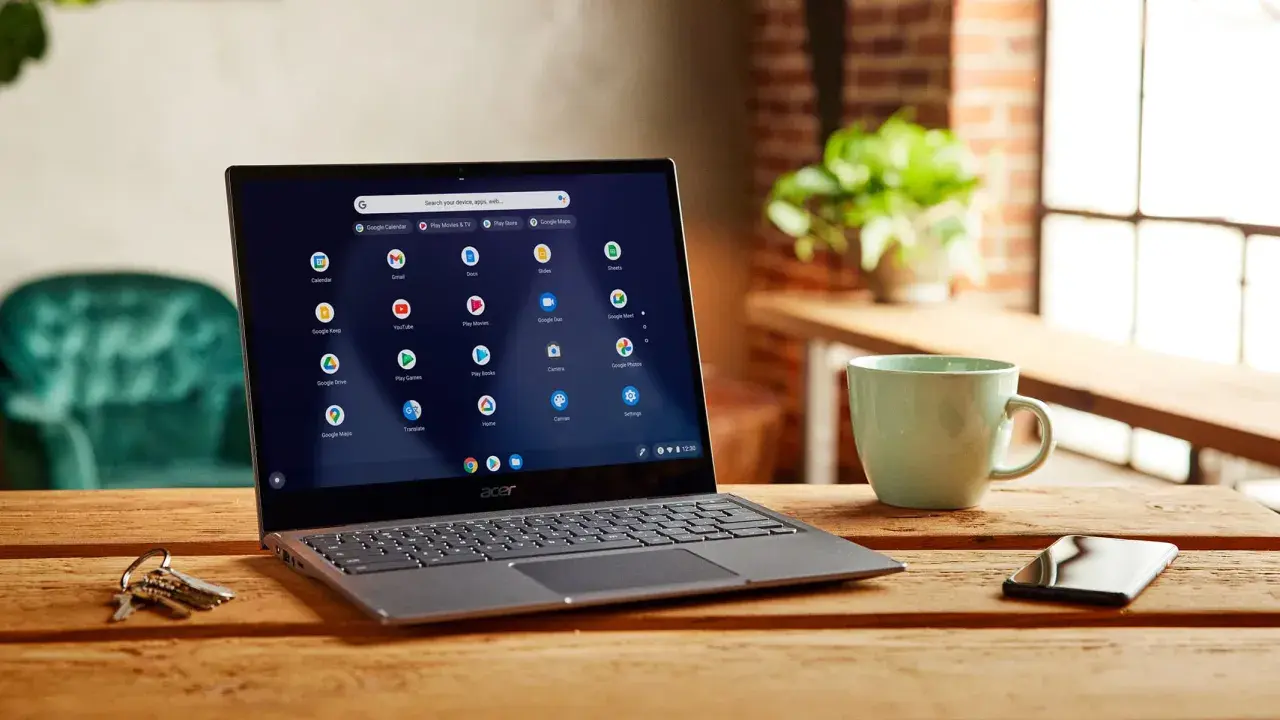 Laptop z systemem Chrome OS instalacja. Na ekranie widoczne ikony aplikacji Google. Obok kubek i klucze.