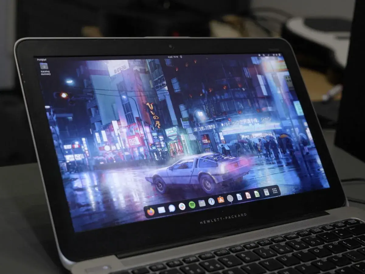 Laptop z systemem Vanilla OS, na ekranie futurystyczne miasto nocą, deszcz i samochód DeLorean.