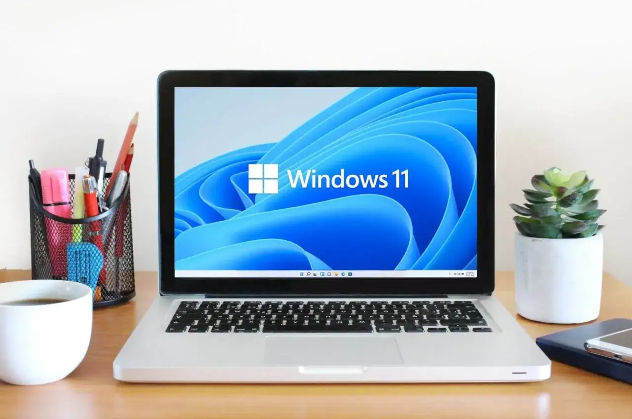 Laptop z ekranem Windows 11, idealny do poradnika jak zainstalować Windows 11 na starym komputerze. Obok kubek kawy i przybory biurowe.